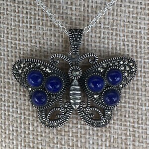 May include: Silver-toned butterfly pendant necklace with a delicate chain. The butterfly design features intricate detailing and is adorned with round, deep blue gemstones. The wings are embellished with small, sparkling accents, creating a beautiful and eye-catching piece of jewelry.