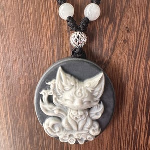 May include: A necklace featuring a round, dark gray pendant with a carved white fox design. The necklace has a black cord, white beads, and a silver-colored decorative bead.