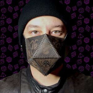 Pode incluir: Uma pessoa usando um gorro preto e uma máscara facial de couro marrom com um design geométrico. O fundo é um padrão preto e roxo de dados.