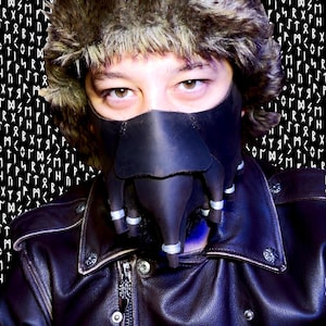 Puede incluir: Una persona con una chaqueta de cuero negro, una máscara de cuero negro con superficie texturizada y un gorro de piel marrón. Los ojos de la persona son visibles a través de la máscara.