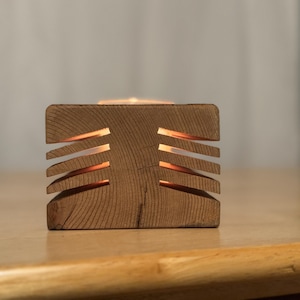May include: A wooden candle holder with a geometric design. The holder has a rectangular base and a series of horizontal slots cut into the sides. A candle is burning inside the holder, casting a warm glow.