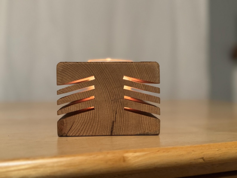 May include: A wooden candle holder with a geometric design. The holder has a rectangular base and a series of horizontal slots cut into the sides. A candle is burning inside the holder, casting a warm glow.