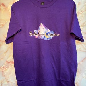 Puede incluir: Una camiseta morada con un dise&ntilde;o gr&aacute;fico que presenta cristales, una luna creciente y el texto "Bella Luna Cosmetics". La camiseta se muestra en una percha blanca.