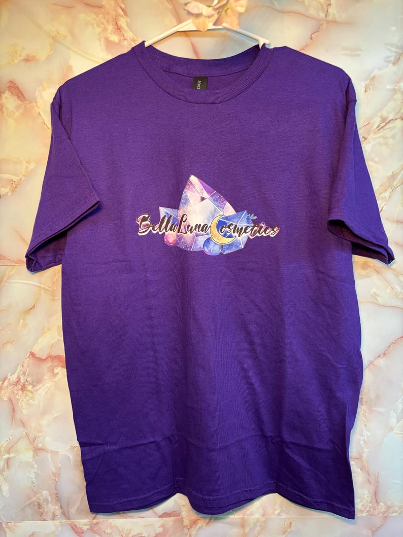 Puede incluir: Una camiseta morada con un dise&ntilde;o gr&aacute;fico que presenta cristales, una luna creciente y el texto "Bella Luna Cosmetics". La camiseta se muestra en una percha blanca.