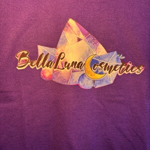 Puede incluir: Una camiseta morada con el logo de Bella Luna Cosmetics. El logo incluye el texto "Bella Luna Cosmetics" en oro, un dise&ntilde;o geom&eacute;trico colorido y una luna creciente.