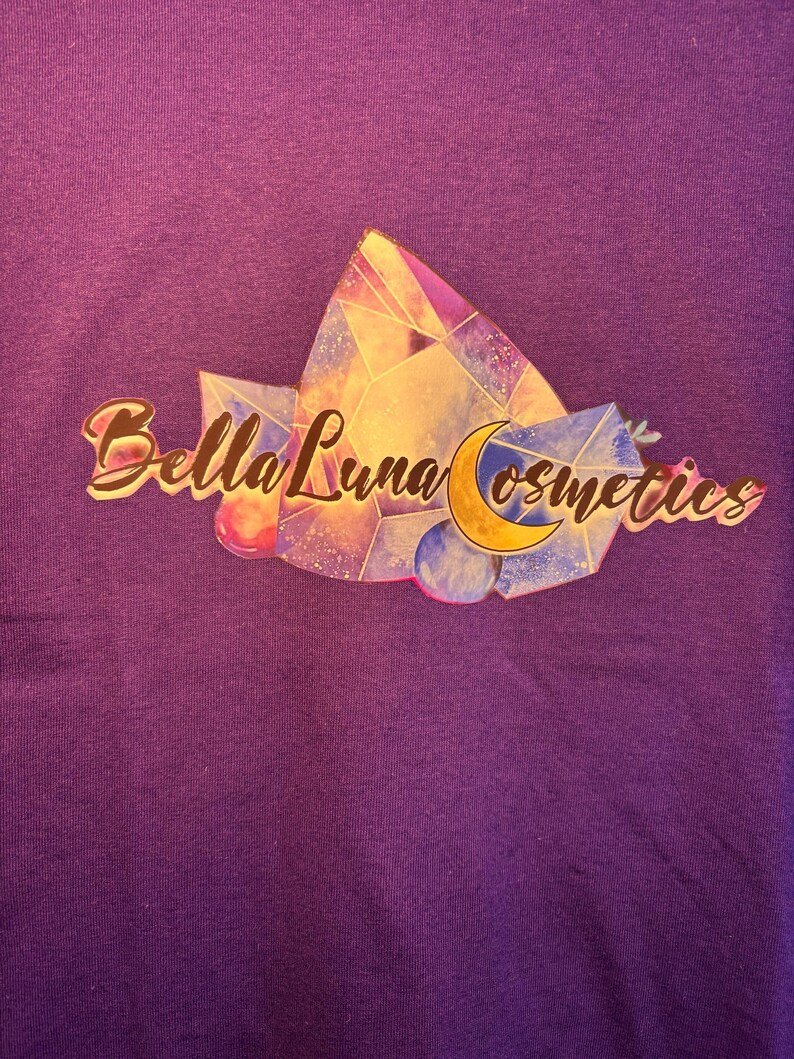 Puede incluir: Una camiseta morada con el logo de Bella Luna Cosmetics. El logo incluye el texto "Bella Luna Cosmetics" en oro, un dise&ntilde;o geom&eacute;trico colorido y una luna creciente.