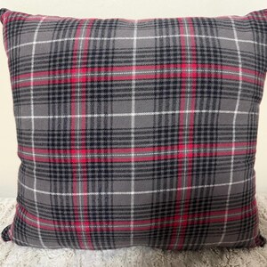 Puede incluir: Cojín decorativo cuadrado con un patrón de cuadros gris, negro, blanco y rojo. El cojín tiene un aspecto suave y texturizado y probablemente está hecho de tela. El diseño de cuadros presenta líneas que se cruzan.