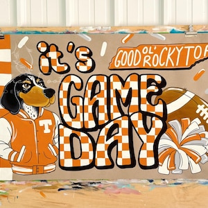 May include: A brown and tan illustration on a tan background with the words "It's Game Day" in orange and white checkered letters. A cartoon dog in an orange jacket and a football are also featured. The words "Good Ol' Rocky Top" are written above.