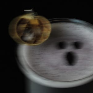 May include: A blurred image of a Halloween-themed design. A small, pumpkin-shaped ornament hangs near a white, circular object with a ghost-like face. The background is solid black.