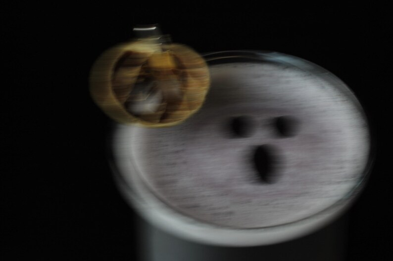May include: A blurred image of a Halloween-themed design. A small, pumpkin-shaped ornament hangs near a white, circular object with a ghost-like face. The background is solid black.