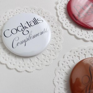 Peut inclure: Bouton blanc avec le texte "Cocktails Compliments" en script noir, placé sur un napperon en dentelle blanc. D'autres boutons sont visibles, dont un bouton rouge avec le texte "Poor Decisions".