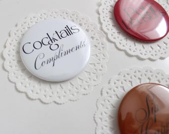 May include: White button with the text "Cocktails Compliments" in black script, placed on a white lace doily. Other buttons with text are visible, including a red button with the text "Poor Decisions".