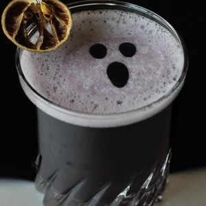 May include: A Halloween-themed cocktail in a clear glass. The drink is dark gray with a white foamy top, featuring a ghost face design. A dried orange slice garnish sits on the rim. The glass has a decorative, ridged design.
