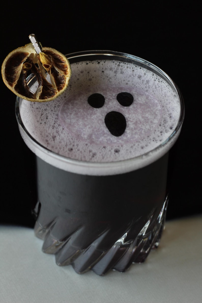 May include: A Halloween-themed cocktail in a clear glass. The drink is dark gray with a white foamy top, featuring a ghost face design. A dried orange slice garnish sits on the rim. The glass has a decorative, ridged design.