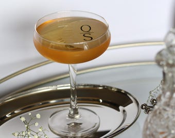May include: A coupe glass filled with a golden-colored liquid, possibly a cocktail, sits on a silver tray. The glass has a long, slender stem and a wide, rounded bowl. The letters "O S" are printed on the glass. A decorative bottle is in the background.