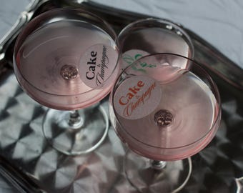 May include: Three stemmed champagne glasses filled with a pink liquid, possibly a cocktail or champagne. Each glass has a circular sticker that reads "Cake & Champagne." The glasses sit on a silver tray, creating a celebratory atmosphere.