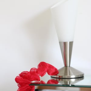 Könnte beinhalten: Eine Tischleuchte mit konischem weißem Glasschirm und einem gebürsteten silbernen Metallsockel, begleitet von einem langen, gerüschten roten Organzaband, das sich in einem Spiralmuster über die Fläche vor dem Lampensockel windet.
