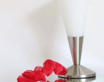 May include: A table lamp with a conical white glass shade and a brushed silver metal base, accompanied by a long, ruffled red organza ribbon that curves in a spiral pattern across the surface in front of the lamp base.