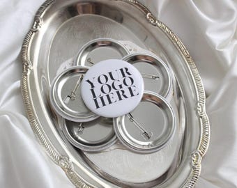 May include: A collection of silver-colored pin-back buttons, one with the text "YOUR LOGO HERE" in black, arranged on a silver oval tray. The tray has a decorative edge and sits on a white, textured fabric.