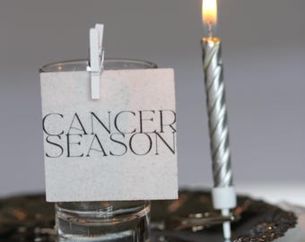 May include: A lit silver birthday candle next to a small glass with a white card that reads "CANCER SEASON" clipped to it. The candle and glass sit on a decorative silver plate.
