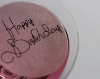 May include: A close-up shot of a pink, circular object with the words "Happy Birthday" written in black cursive script. The object is inside a clear glass, with a white background.