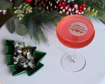 May include: A festive cocktail in a stemmed glass, topped with a "Merry Christmas" sticker. A green, tree-shaped dish holds wrapped chocolates. The scene is decorated with pine and red berries, creating a holiday theme.