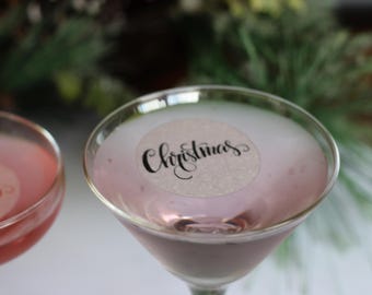 May include: Two martini glasses filled with a pink liquid, one with a sticker that says "Christmas" in elegant script. The glasses are on a white surface, with greenery in the background.