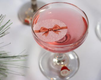 May include: A pink cocktail in a coupe glass, garnished with a white, round edible decoration featuring a brown bow. The glass has a decorative charm. Festive holiday drink.