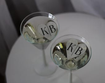 May include: Two martini glasses filled with a clear liquid, each with a heart-shaped decoration bearing the initials "KB" in black. The glasses sit on a white surface, suggesting a celebratory occasion.