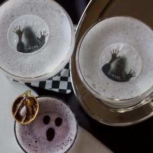 May include: Overhead shot of three cocktails, two with a spooky design and the word "Cheers" on the foam. The third cocktail has a ghost face design and a dried orange slice. The drinks are on a silver tray and a black and white checkered napkin.