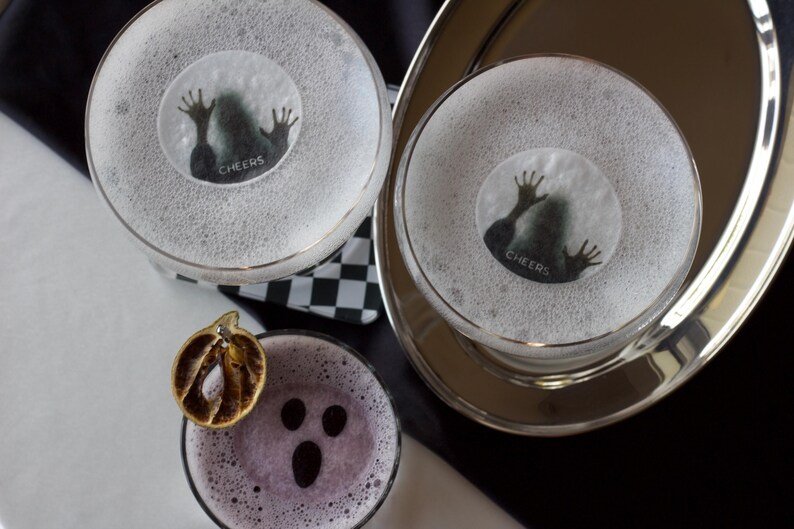 May include: Overhead shot of three cocktails, two with a spooky design and the word "Cheers" on the foam. The third cocktail has a ghost face design and a dried orange slice. The drinks are on a silver tray and a black and white checkered napkin.