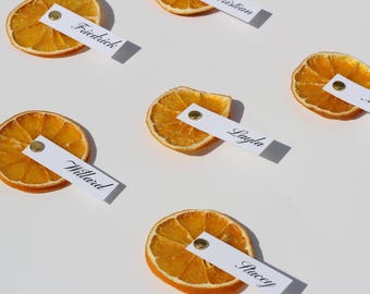May include: Dried orange slices with white name tags attached with gold brads. The name tags are printed with names in a cursive font. The orange slices are arranged on a white surface.