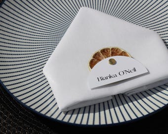 May include: A white folded napkin with a dried orange slice and a name tag that reads "Bianka O'Neil" rests on a blue and white striped plate. The plate is on a dark textured surface, suggesting a place setting.
