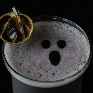 May include: A Halloween-themed cocktail in a clear glass, featuring a foamy, light purple drink with a ghost face design made of black circles. Garnished with a dried orange slice and a metal pick. Set against a black background.