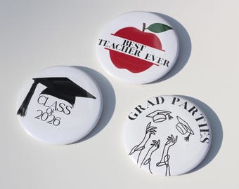 May include: Three white round buttons with black and red graphics. One button has a red apple with the text "BEST TEACHER EVER". Another has a graduation cap and the text "CLASS OF 2026". The third has the text "GRAD PARTIES" and graduation caps.