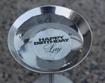 May include: A silver metal candle holder with a round top. A circular paper insert with the words "HAPPY BIRTHDAY Lay" in black lettering is centered on the top.