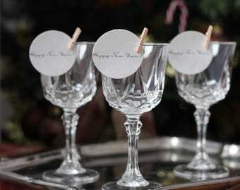 May include: Three crystal wine glasses on a silver tray, each adorned with a small, round tag that reads "Happy New Year" and held in place by a tiny wooden clothespin. The glasses have intricate cut-glass designs.