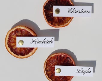 May include: Three dried blood orange slices with white name cards attached with gold brads. The cards read "Christian", "Friedrick", and "Layla" in elegant script. The arrangement is on a white background.