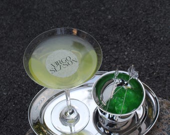 May include: A cocktail set on a silver tray. A martini glass with a yellow-green drink and a round label that says "Virgo Season". Next to it is a small silver bucket with a green drink and a strainer.