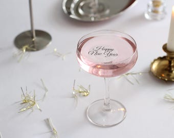May include: A cocktail glass filled with a pink liquid, with the words "Happy New Year" printed on the surface. A lit candle in a gold candlestick and decorative accents are also visible, creating a festive atmosphere.