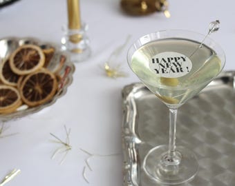 May include: A martini glass with a Happy New Year message on a white circle, garnished with an olive. The drink sits on a silver tray, with dried orange slices in a silver dish nearby. Festive New Year's Eve theme.