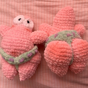 May include: Two handmade crochet plush toys shaped like a pink starfish character. Each figure features a textured yarn surface, a light green waistband with small purple accents, and one figure displays two black eyes on a lighter pink face.