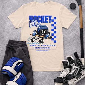 Op de afbeelding: Cr&egrave;mekleurig T-shirt met een blauw-zwarte hockey-afbeelding. Het shirt toont een cartoon hockeyspeler en de woorden "HOCKEY Vibes". Ook afgebeeld zijn hockeyschaatsen, een hockeystick, een puck, een joggingbroek en hockeyhandschoenen.