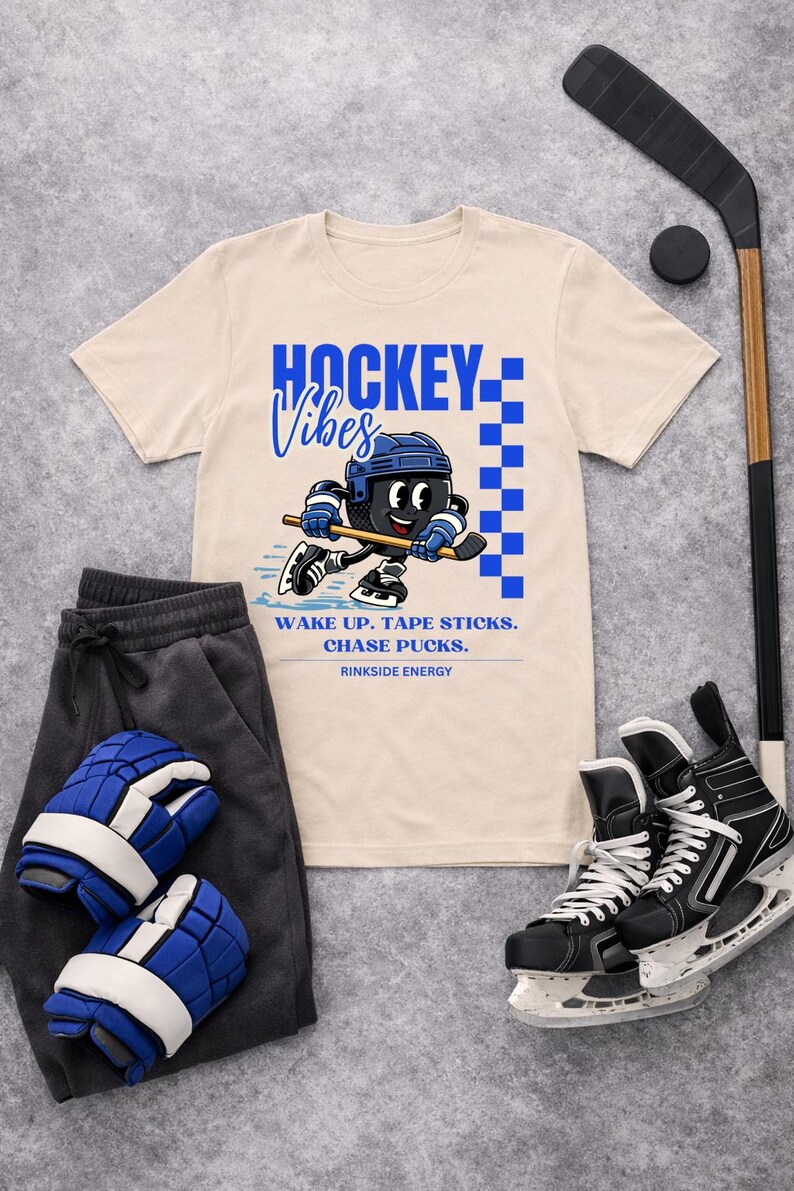 Op de afbeelding: Cr&egrave;mekleurig T-shirt met een blauw-zwarte hockey-afbeelding. Het shirt toont een cartoon hockeyspeler en de woorden "HOCKEY Vibes". Ook afgebeeld zijn hockeyschaatsen, een hockeystick, een puck, een joggingbroek en hockeyhandschoenen.