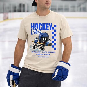 Op de afbeelding: Beige t-shirt met blauwe "HOCKEY Vibes" tekst en een cartoon hockey speler graphic. Het shirt heeft ook de tekst "WAKE UP. TAPE STICKS. CHASE PUCKS. RINKSIDE ENERGY."