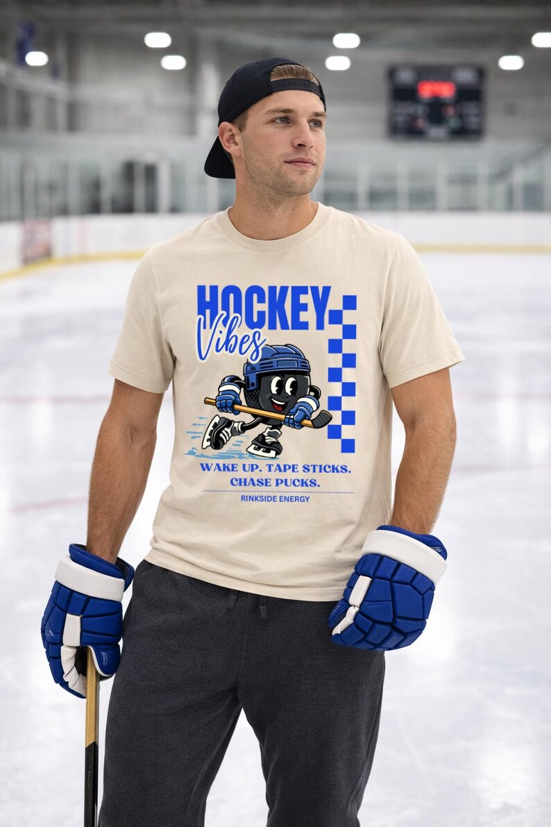 Op de afbeelding: Beige t-shirt met blauwe "HOCKEY Vibes" tekst en een cartoon hockey speler graphic. Het shirt heeft ook de tekst "WAKE UP. TAPE STICKS. CHASE PUCKS. RINKSIDE ENERGY."