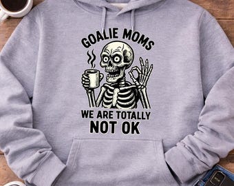 Op de afbeelding: Een lichtgrijze hoodie met een voorzak en een capuchon met trekkoord. De hoodie heeft een afbeelding van een skelet met een koffiekopje met de tekst "GOALIE MOMS WE ARE TOTALLY NOT OK".