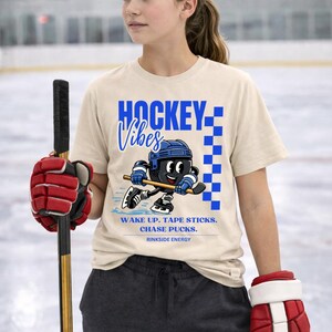 Op de afbeelding: Beige t-shirt met de tekst "HOCKEY Vibes" in blauw en een cartoon hockeyspeler. Het shirt heeft ook de tekst "WAKE UP. TAPE STICKS. CHASE PUCKS. RINK SIDE ENERGY."