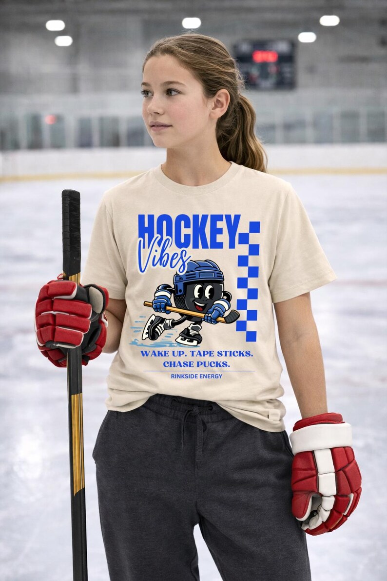 Op de afbeelding: Beige t-shirt met de tekst "HOCKEY Vibes" in blauw en een cartoon hockeyspeler. Het shirt heeft ook de tekst "WAKE UP. TAPE STICKS. CHASE PUCKS. RINK SIDE ENERGY."
