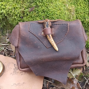 May include: A brown leather pouch with a flap closure, secured by a leather strap and a decorative antler piece. The pouch is set against a backdrop of moss and leaves, suggesting an outdoor or rustic theme. A compass and map are visible.